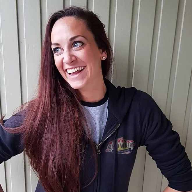 Smiling woman with long brown hair wearing a black hoodie and grey t-shirt against a wooden wall.
