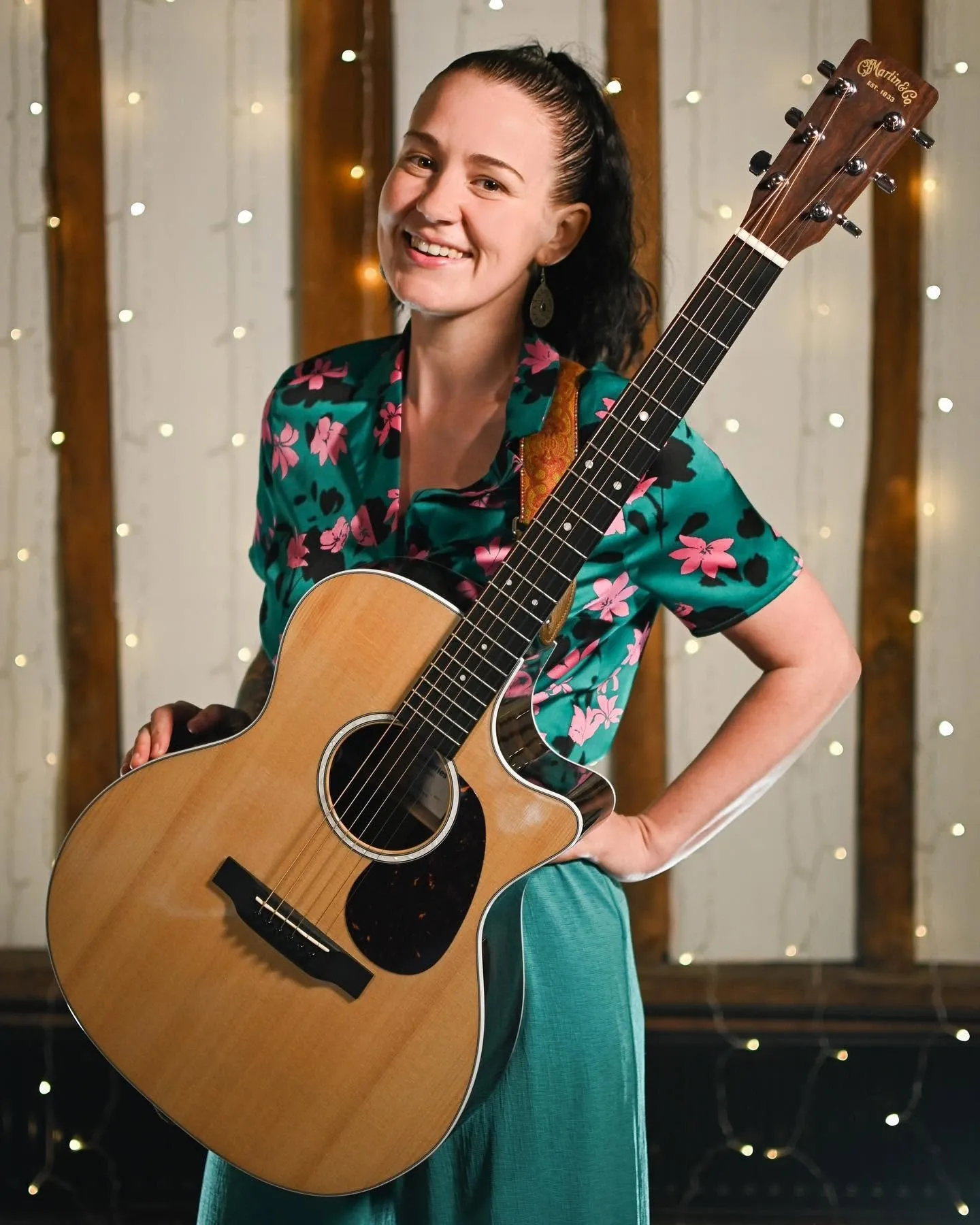 Smiling woman in floral shirt holding an acoustic guitar in a warmly lit room with string lights.