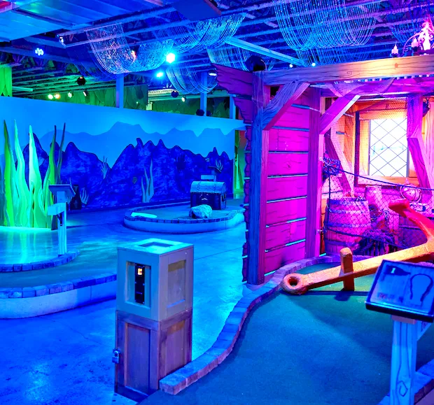 Indoor mini golf course with nautical-themed obstacles and blue and purple lighting.