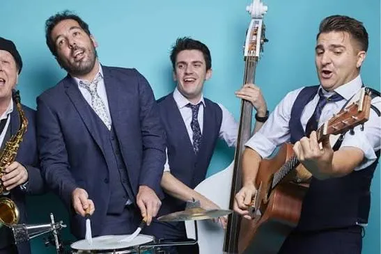 Four male musicians in suits playing drums, saxophone, double bass, and guitar against a blue background.