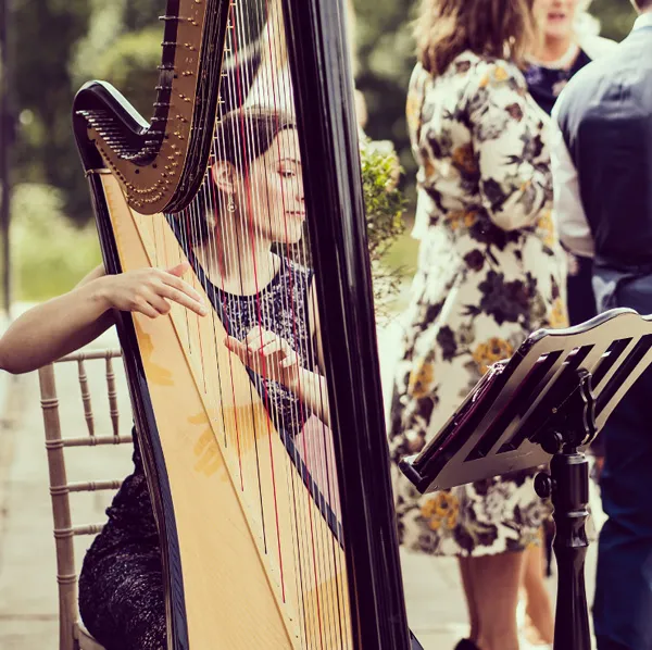 Cecily playing a harp with a sheet music stand nearby, outdoors at a social event.