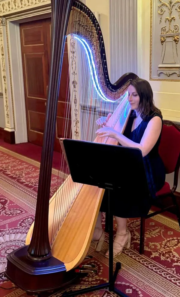 Cecily seated playing a large wooden harp with neon light strings indoors on patterned carpet.