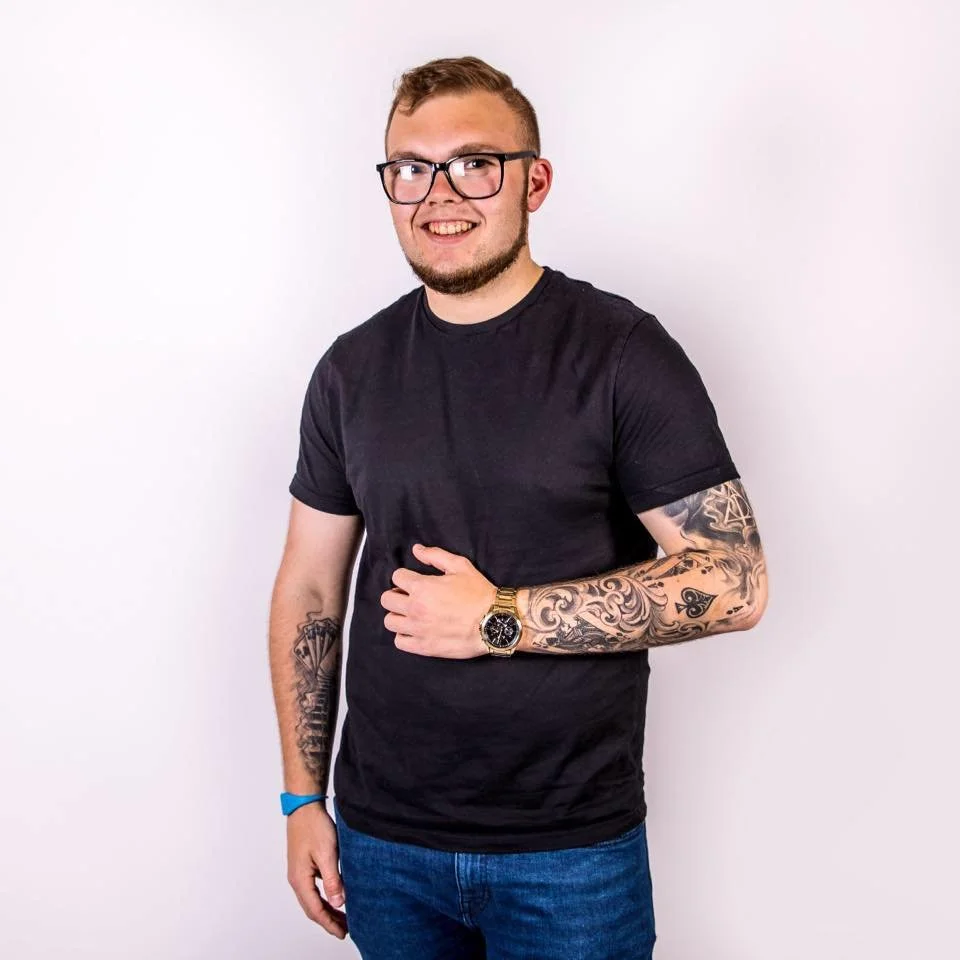 Smiling man with glasses, tattoos on arms, black t-shirt, blue jeans, and gold watch on left wrist.