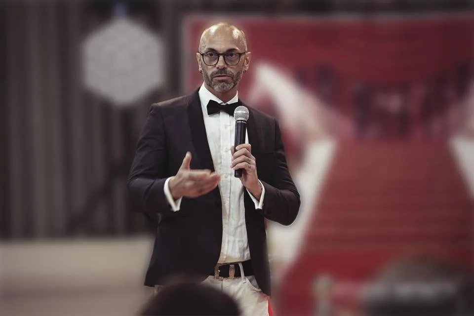 Man in tuxedo and glasses holding microphone giving a performance indoors.