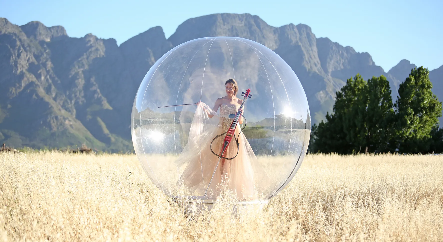Carol in a flowing gown plays a cello inside a large transparent bubble in a field with mountains behind.
