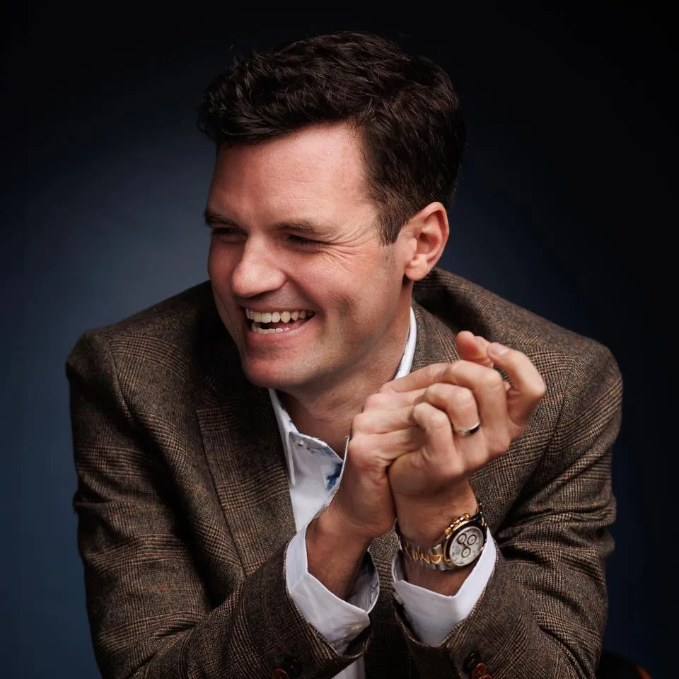 Smiling man in a brown checkered blazer clasping his hands, wearing a wristwatch and wedding ring.