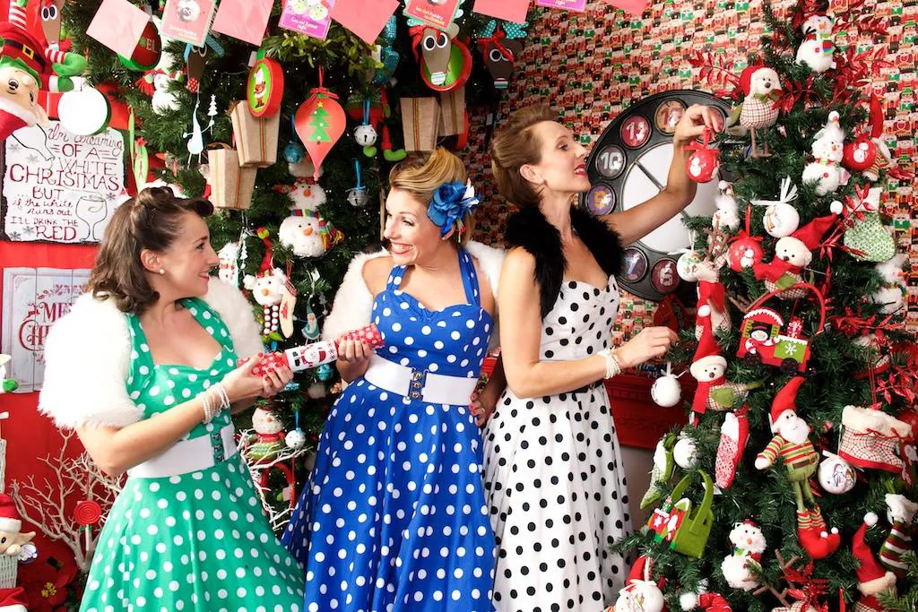 Three women in colorful polka dot dresses decorating a Christmas tree with festive ornaments.
