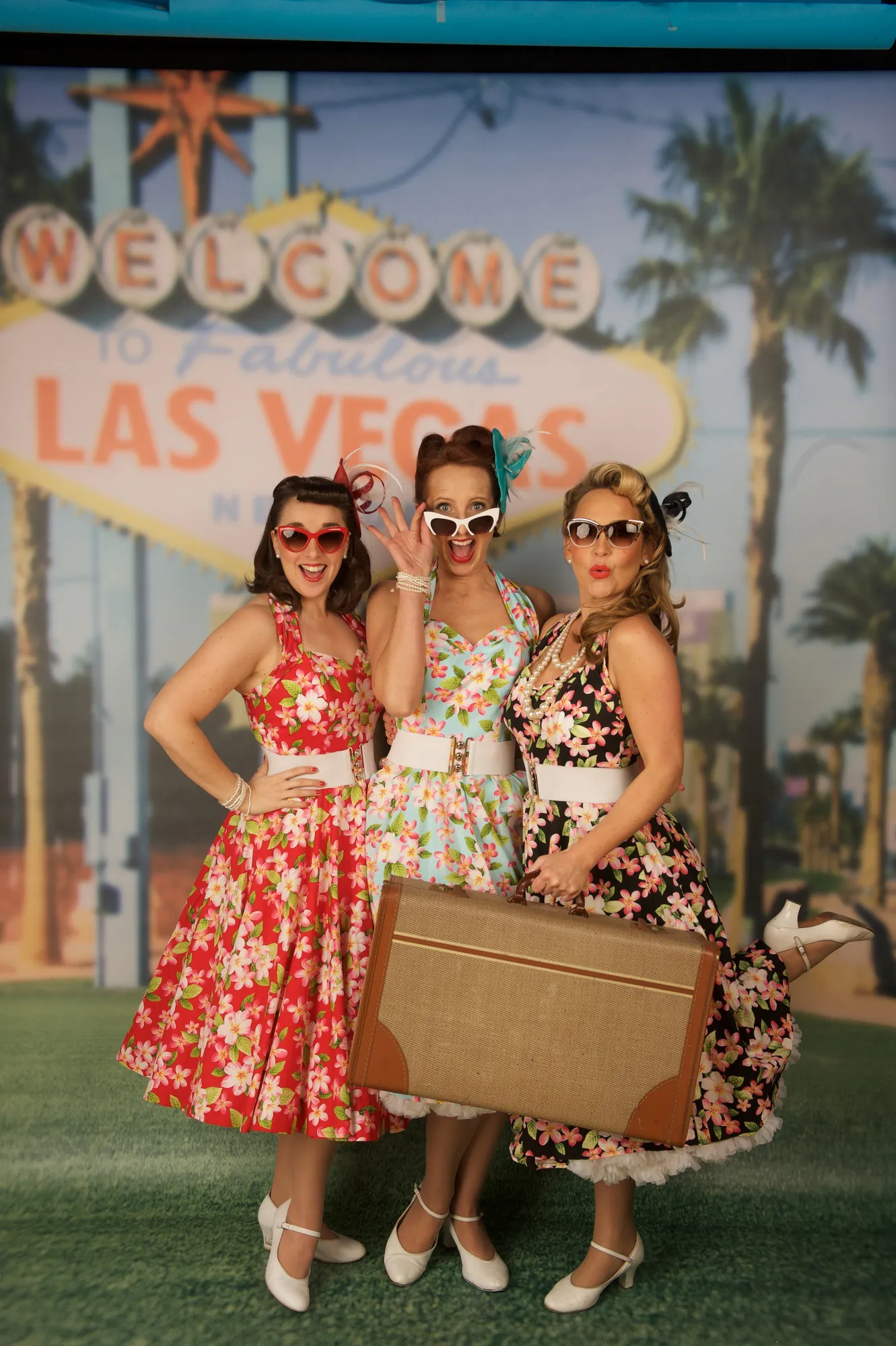 Three women in vintage floral dresses pose with suitcase under Welcome to Fabulous Las Vegas sign.