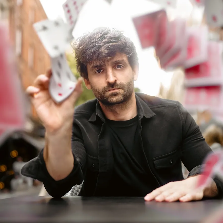 Man with dark hair and beard throwing playing cards toward the camera over a black table.