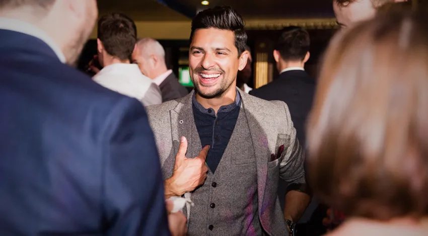Smiling man in gray suit and dark shirt talking with people at a social gathering indoors.