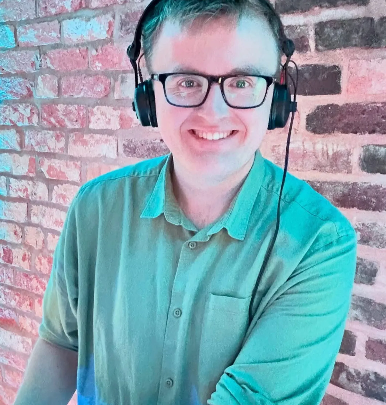 Harry smiling wearing headphones and a green shirt in front of a brick wall.