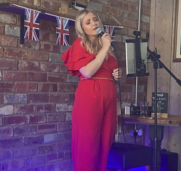 Jessica in red dress singing into a microphone in a room decorated with British flags on a brick wall.