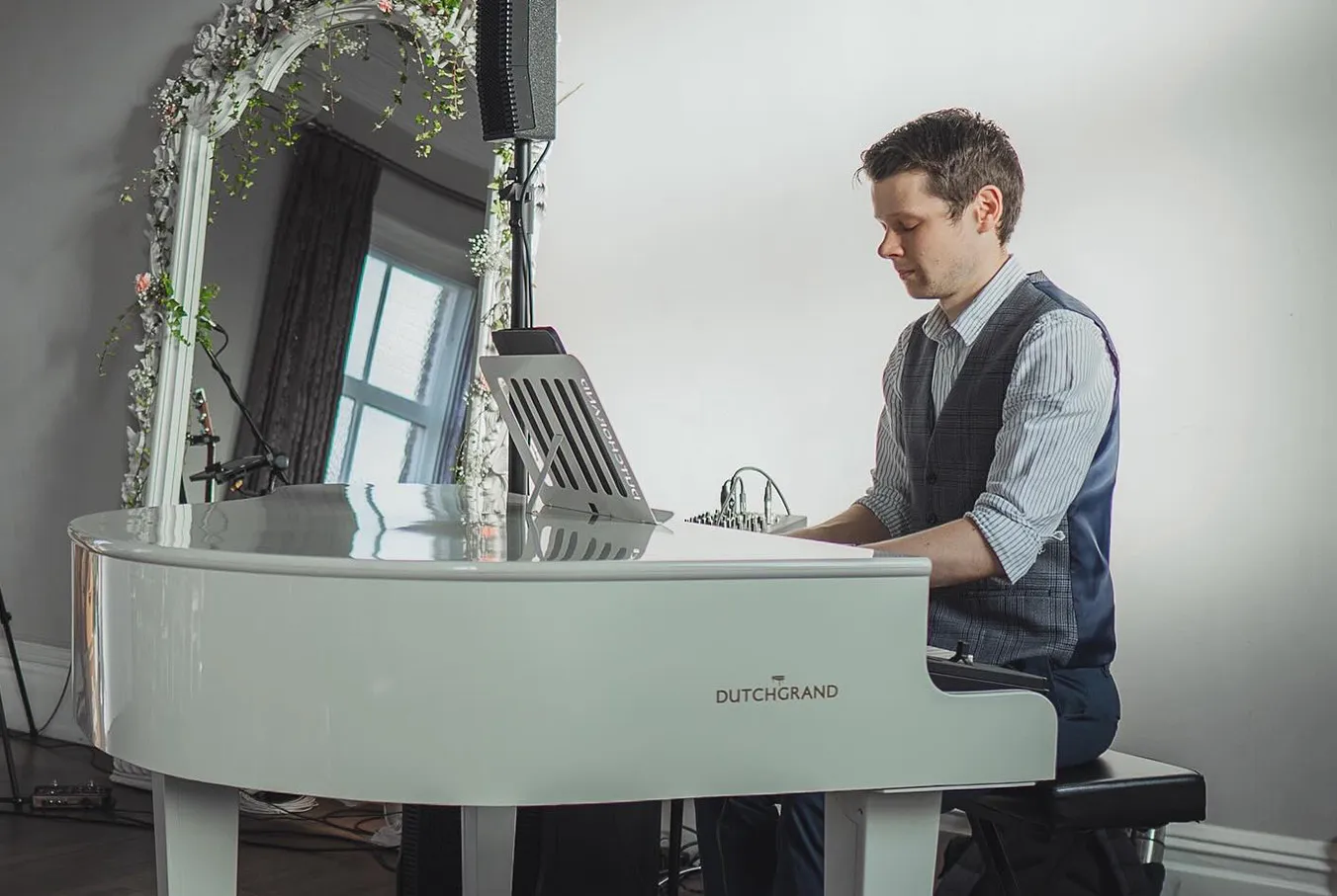 Man in waistcoat playing a white Grand piano in a room with a large decorative mirror.