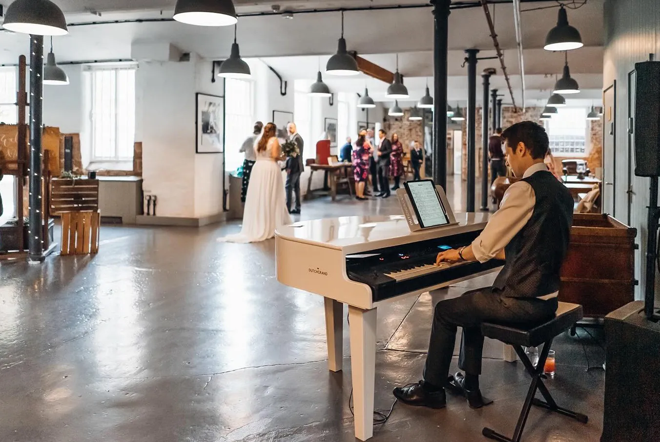 Man in waitscoat playing white piano in a bright event space with people mingling in background.