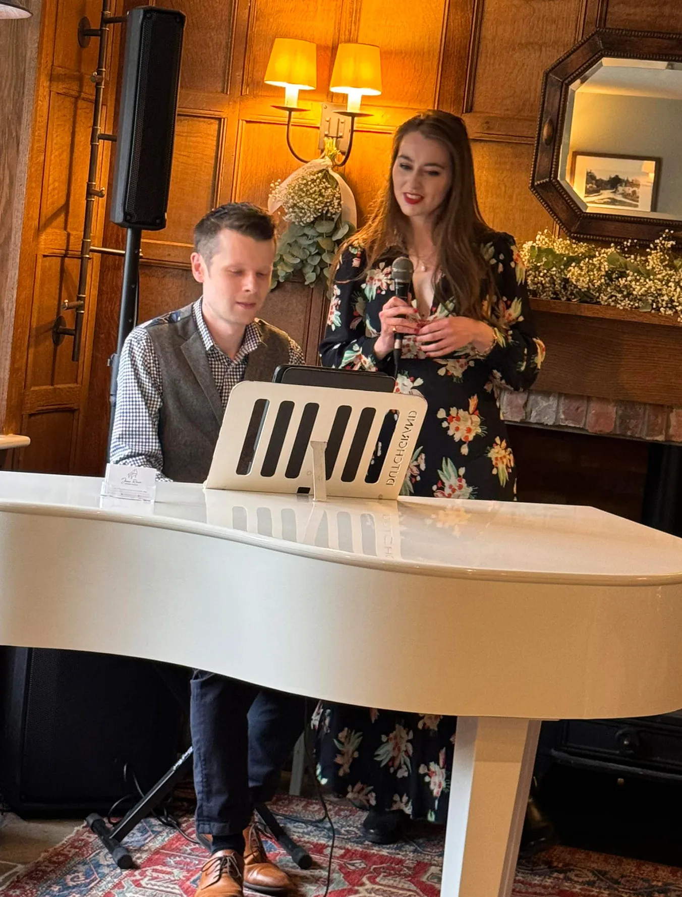 Man playing white grand piano while woman in floral dress sings with microphone in cozy room.