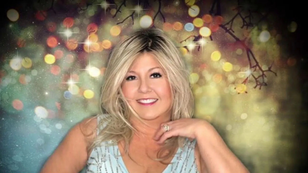 Smiling woman with blonde hair in a light blue dress against a colorful twinkling lights background.