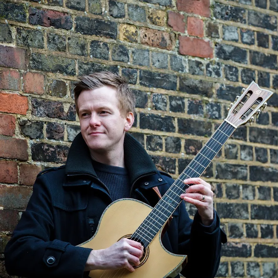 Man playing acoustic guitar while standing against an aged brick wall outdoors.