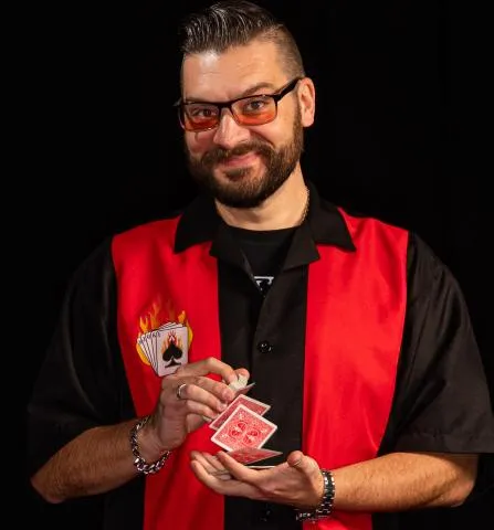 Man with glasses and beard wearing red and black shirt performing card sleight of hand.