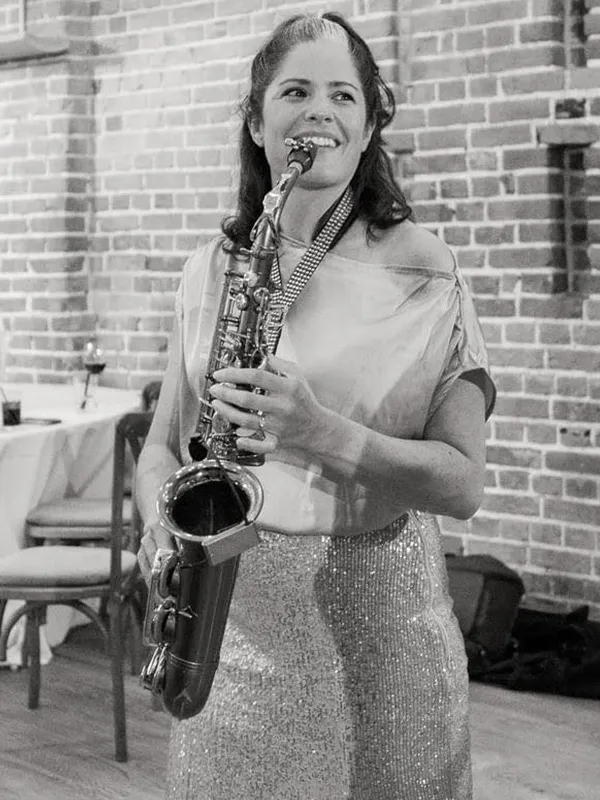 Lucy smiling and holding her saxophone in front of a brick wall and chairs.