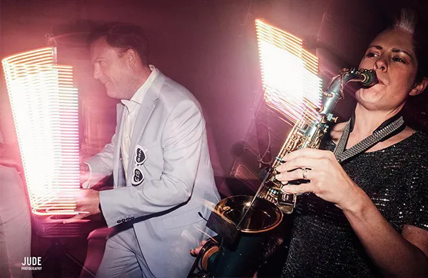 Man in a white suit playing keyboard and Lucy playing saxophone with light trails in the background.