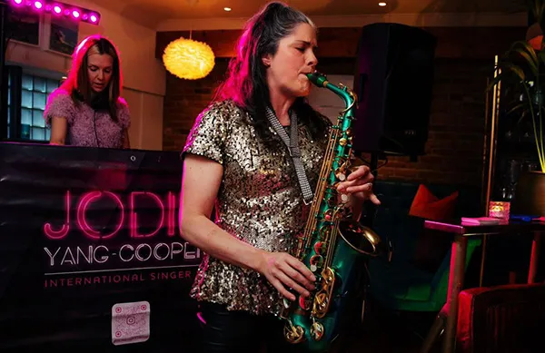 Lucy wearing a shiny gold top playing saxophone in a dimly lit room with a DJ in the background.