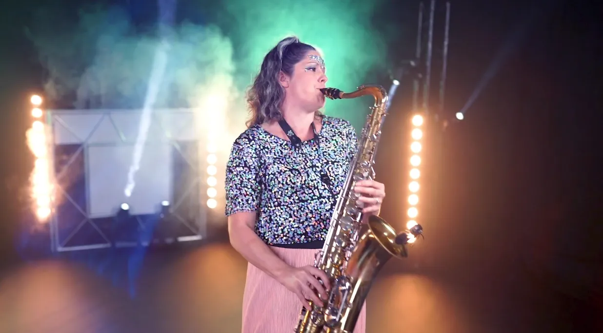 Lucy playing saxophone on stage with colourful lights and smoke in background.