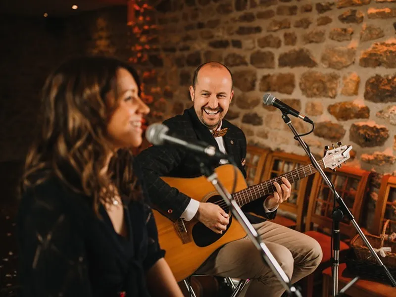 Man playing acoustic guitar and smiling, woman singing into microphone in a cozy brick-walled room.