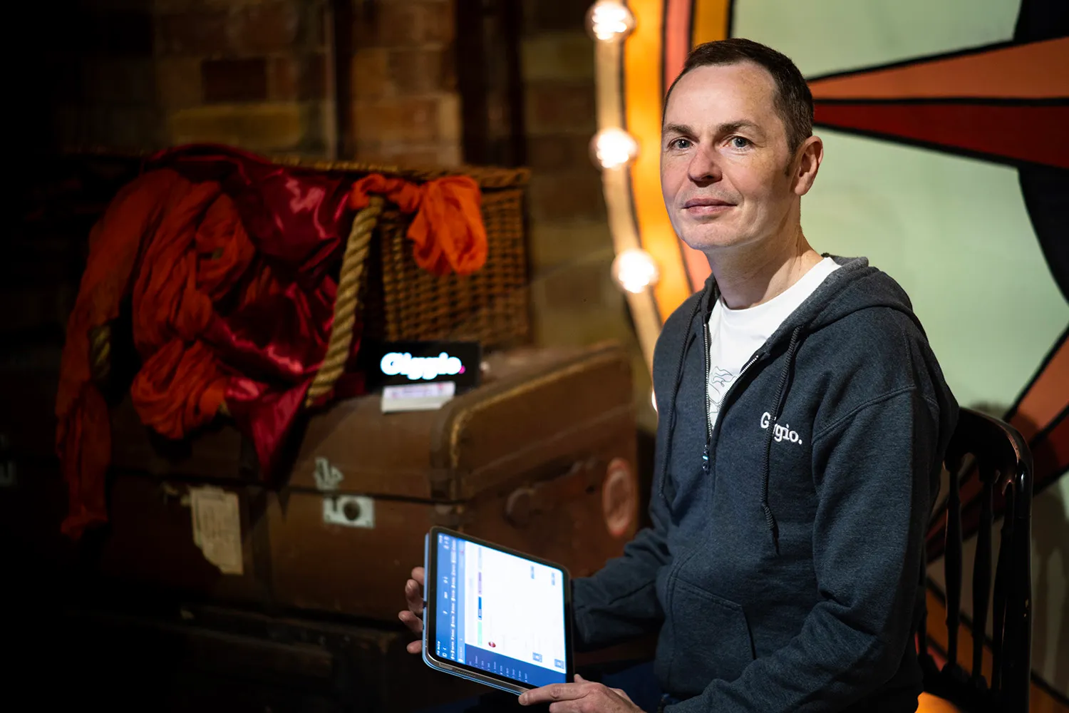 Martyn in Giggio hoodie holding a tablet in a warmly lit room with colourful background.