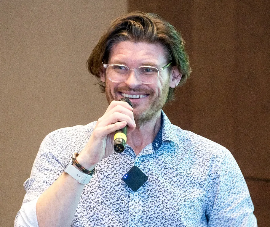 Man with glasses and beard speaking into a microphone wearing a patterned shirt and wristwatch.