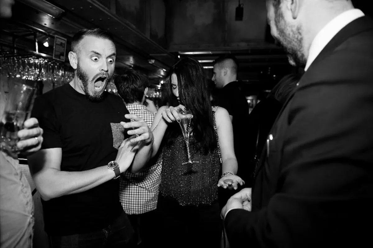 Man reacts with shock as woman spills drink from glass in a crowded bar or party setting.