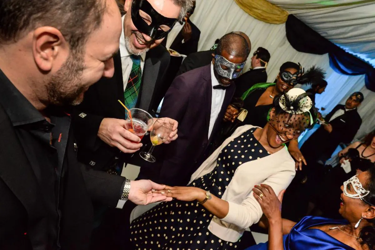 Group of people wearing masquerade masks enjoying a party, with one woman showing her hand and ring.