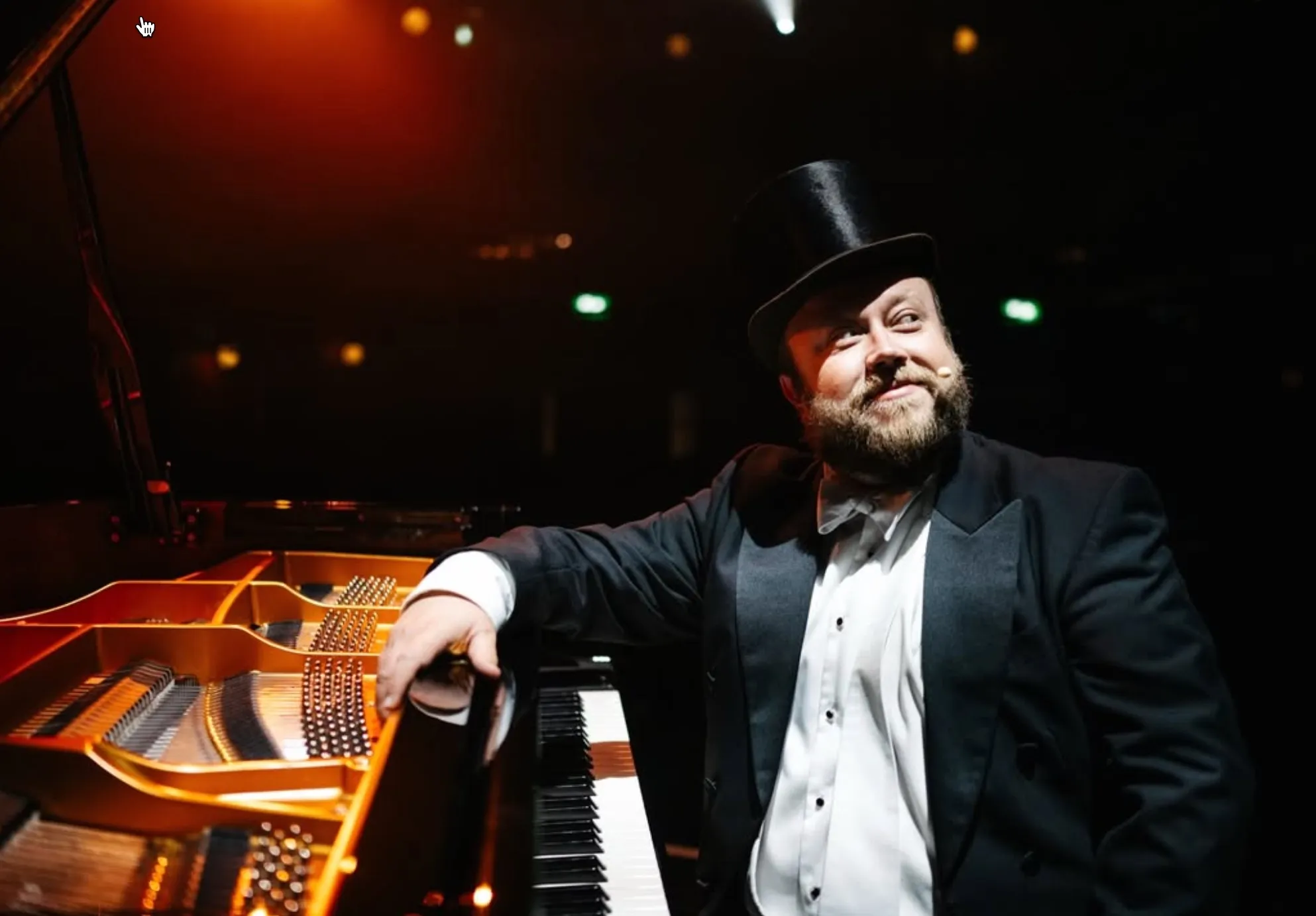Bearded man in tuxedo and top hat near grand piano under warm stage lights.