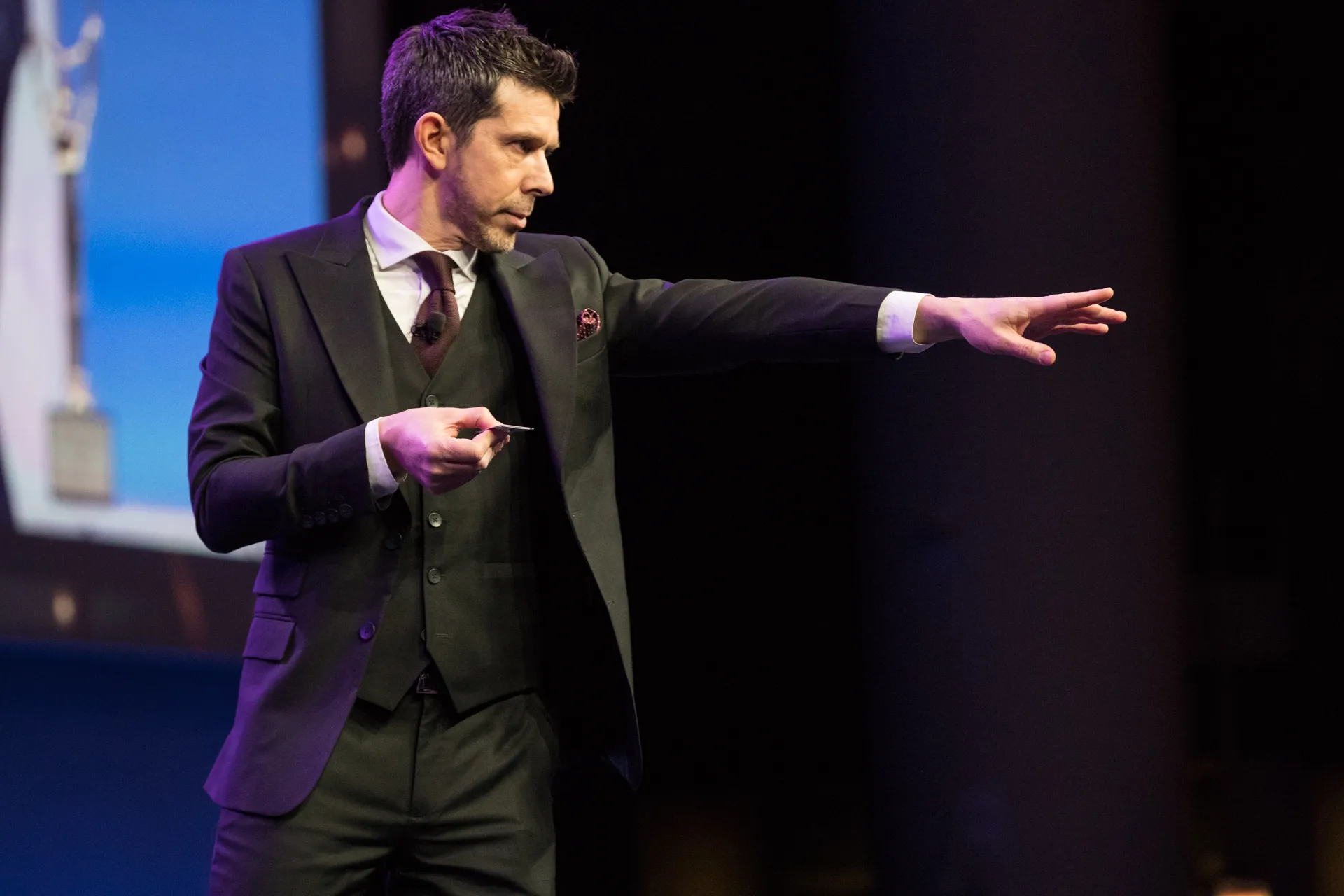 Man in a suit presenting on stage, pointing with his right hand while holding a card in his left.