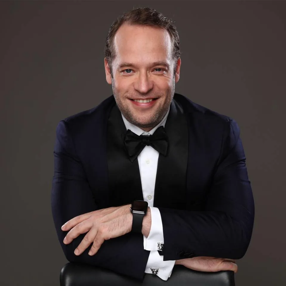 Smiling man in a black tuxedo with bow tie and smartwatch posing with crossed arms against grey background.
