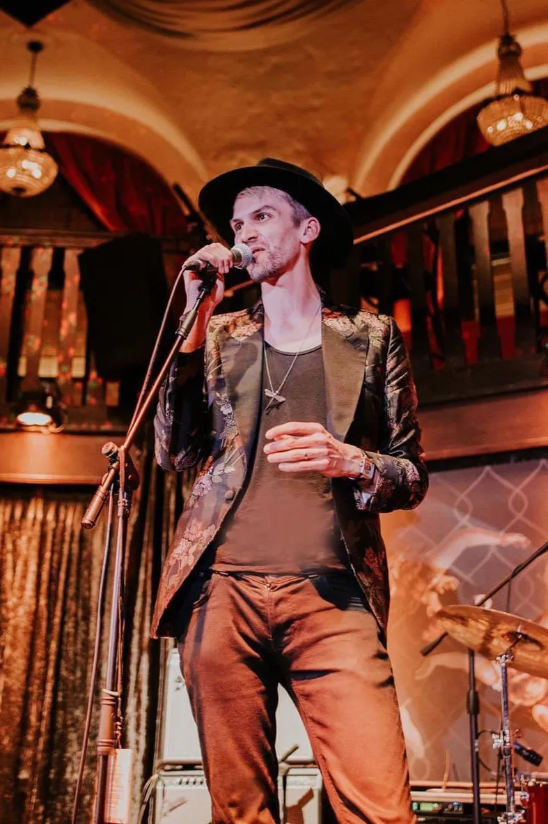 Magician wearing a black hat and floral blazer speaking into a microphone on stage.