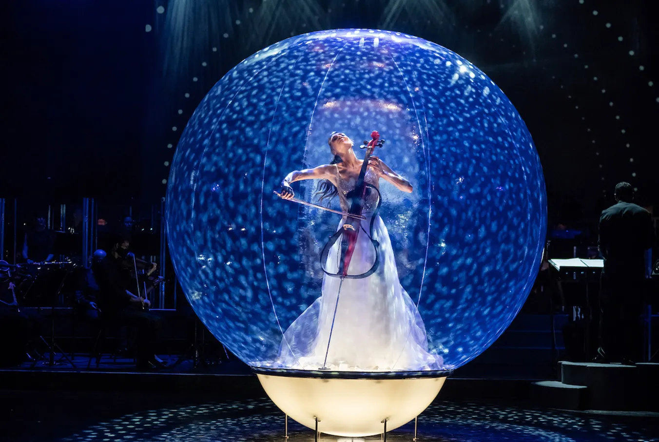 Carol in white dress playing cello inside illuminated blue glass sphere on stage.