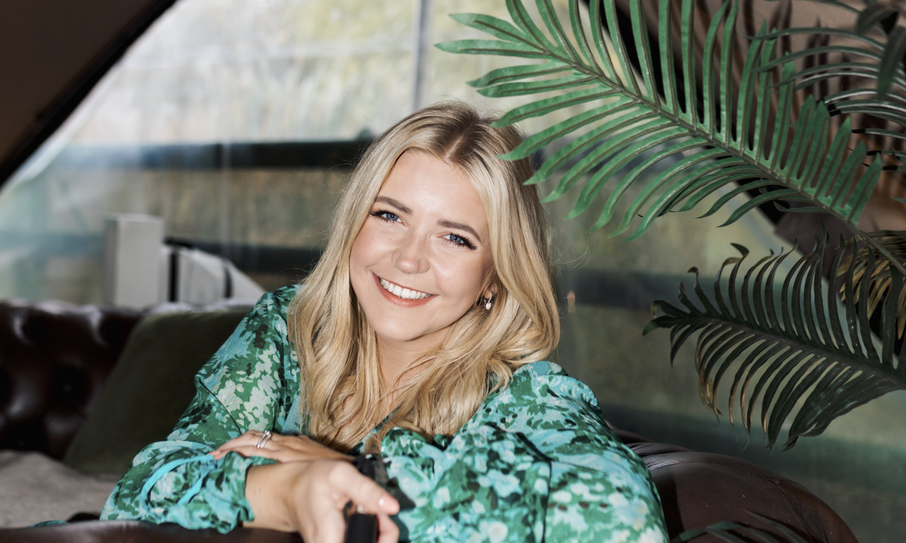 Smiling blonde woman in a green patterned blouse sitting on a leather couch near green plants.