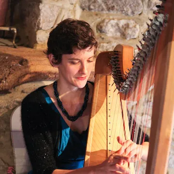 Woman with necklace playing a wooden harp in a warm-lit room with stone walls.