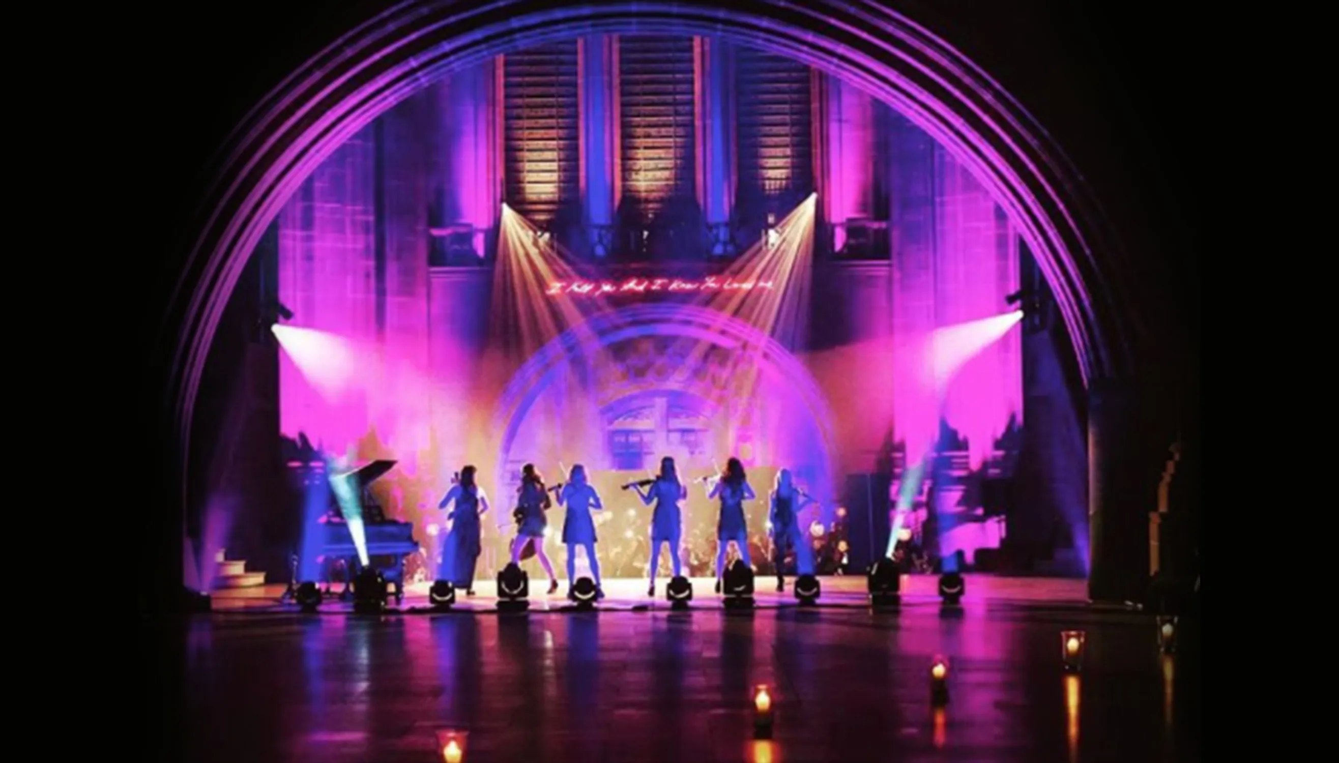 Silhouettes of six musicians performing under purple and pink stage lighting in a large arch venue.