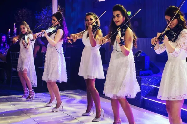 Five musicians in white dresses playing violins on stage under blue lighting.