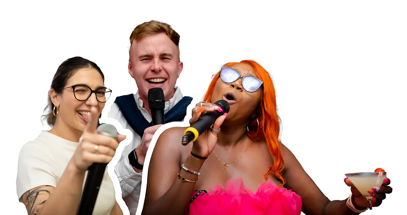 Three people singing with microphones, one holding a cocktail and wearing orange hair and glasses.