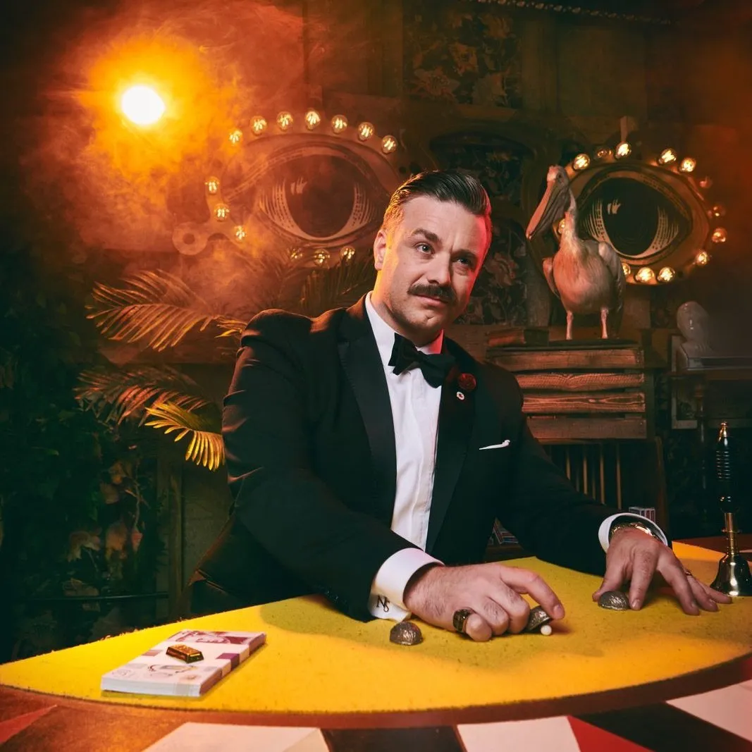 Man in tuxedo with mustache seated at yellow table with cards under warm light in vintage room.