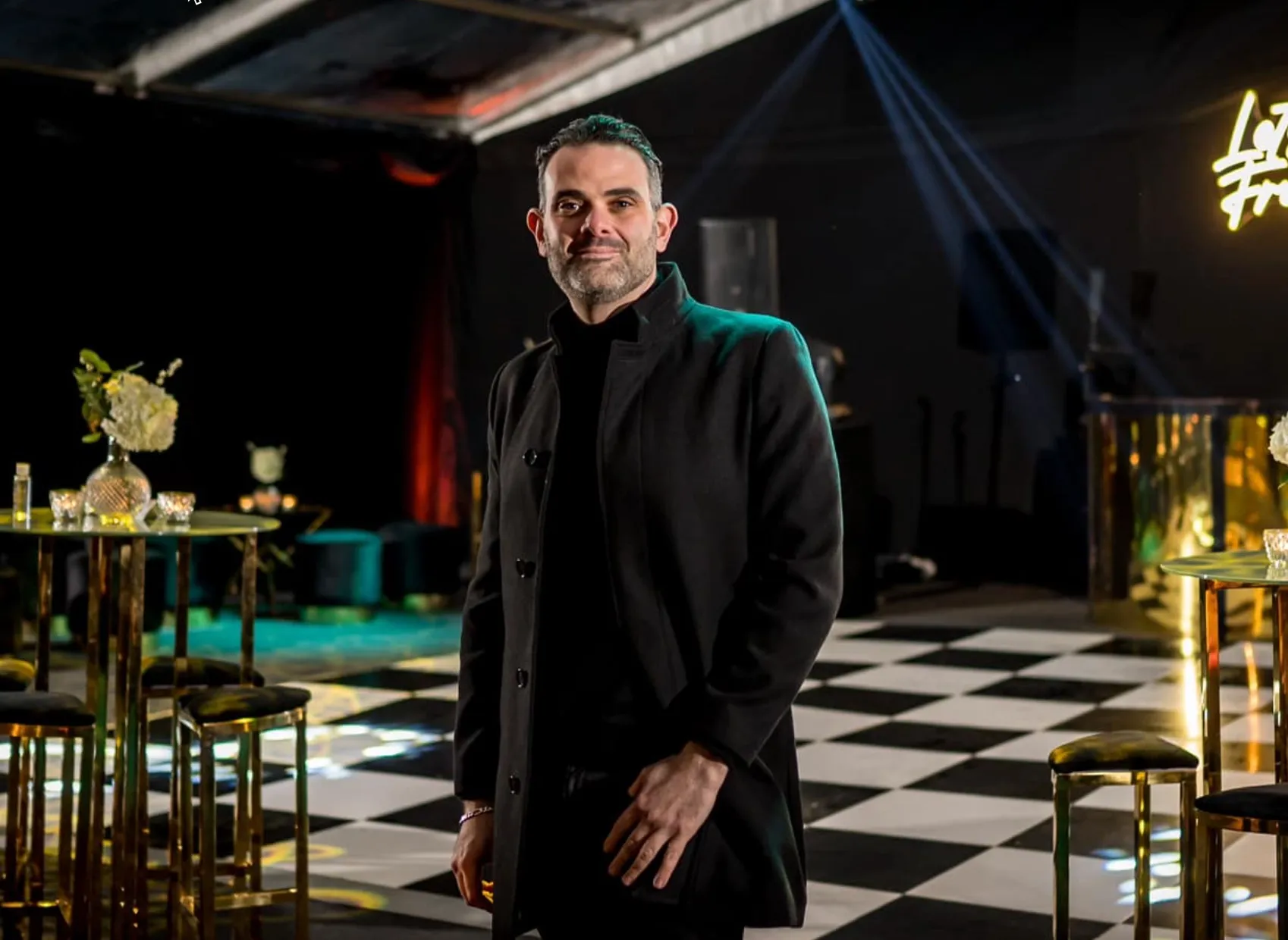 Man in a black coat stands on a black and white checkered floor in a dimly lit event space.