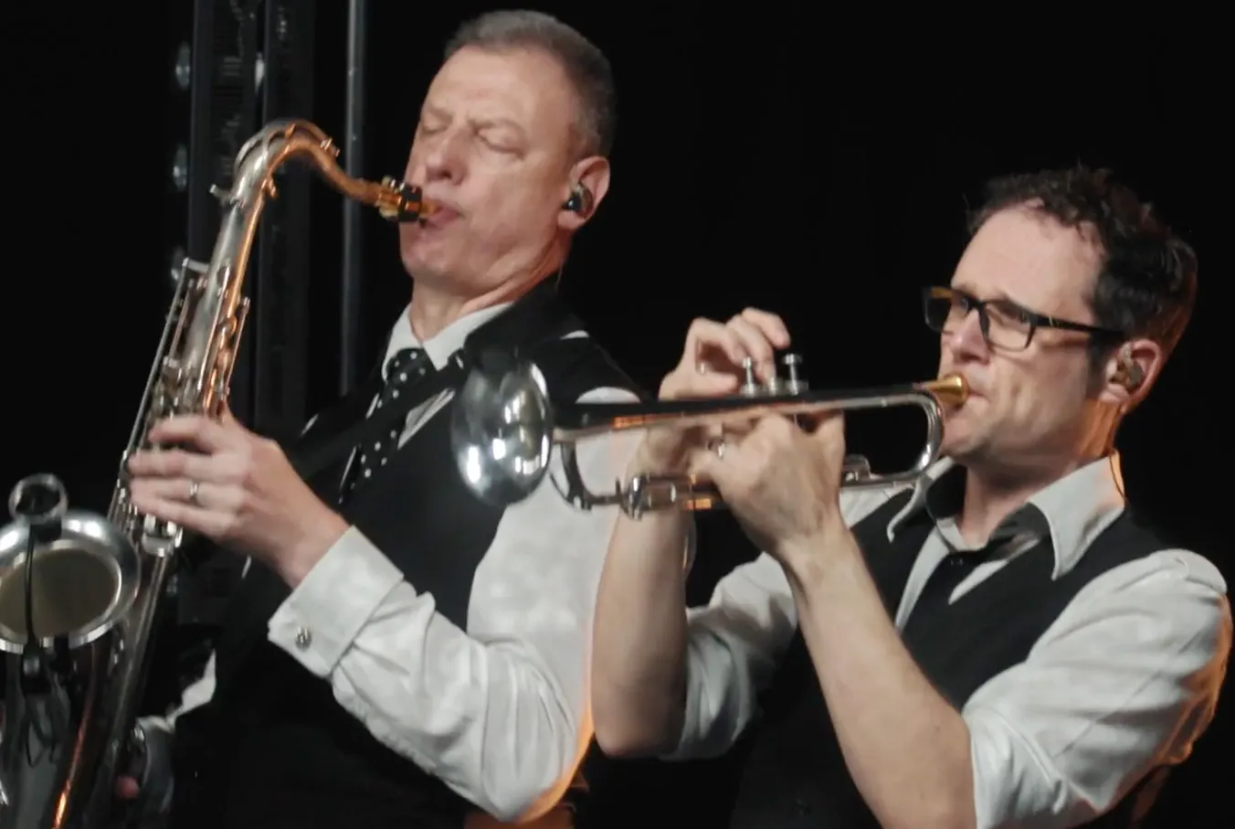 Two men in white shirts and black vests playing saxophone and trumpet with eyes closed.