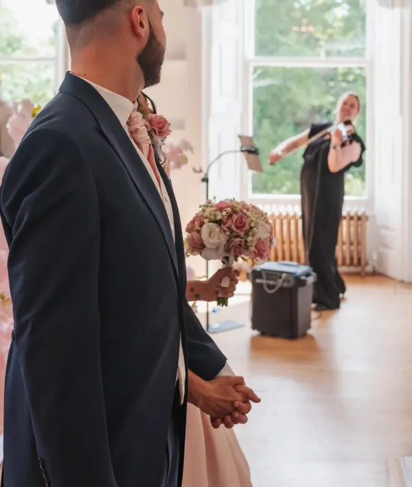 Couple holding hands with bouquet, watching Emily perform in a sunlit room.