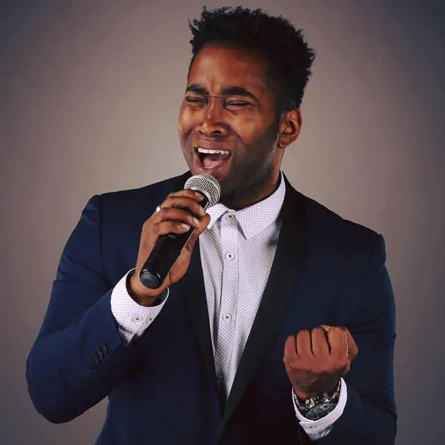 Man in navy blue suit passionately singing into a microphone with eyes closed and fist clenched.