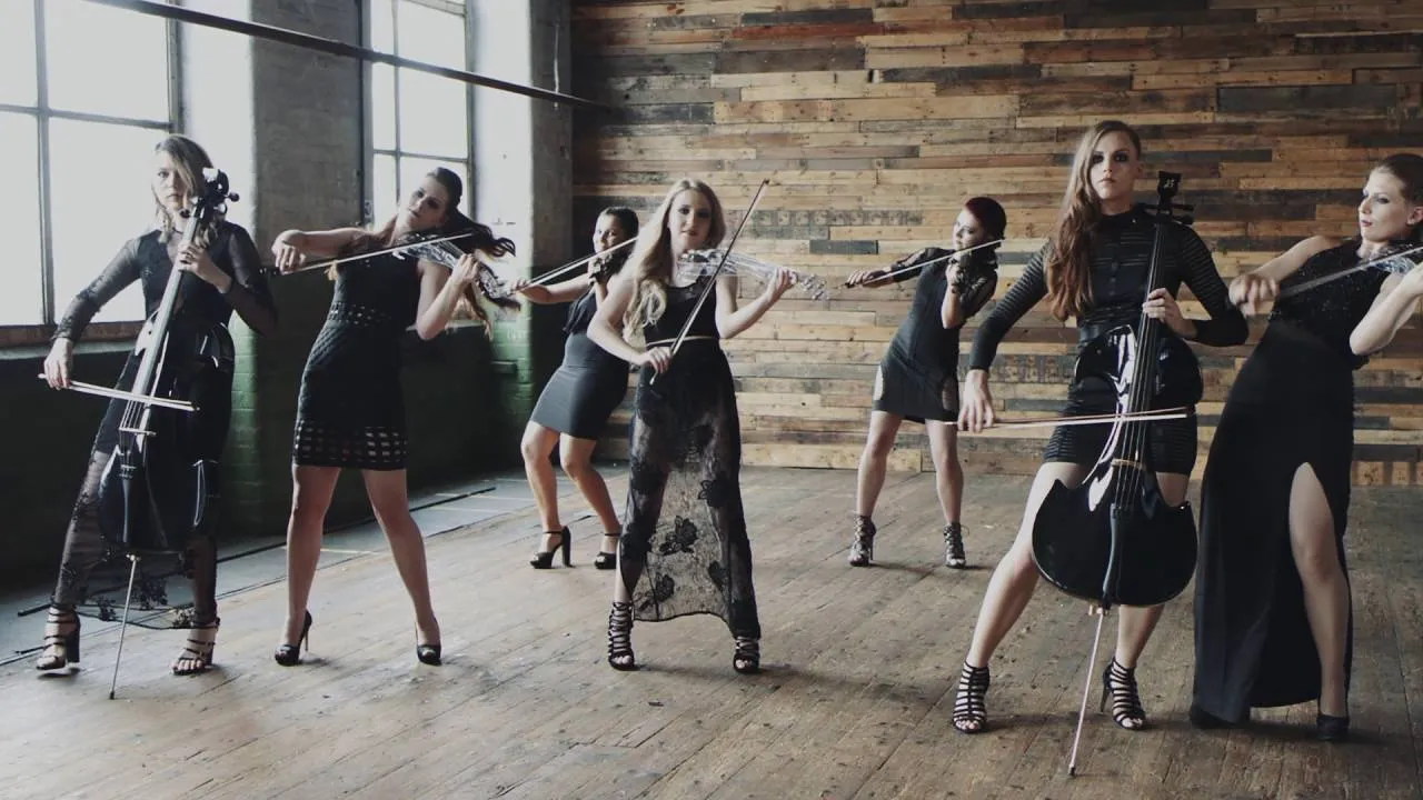 Six women in black dresses playing string instruments in a wooden room with large windows.